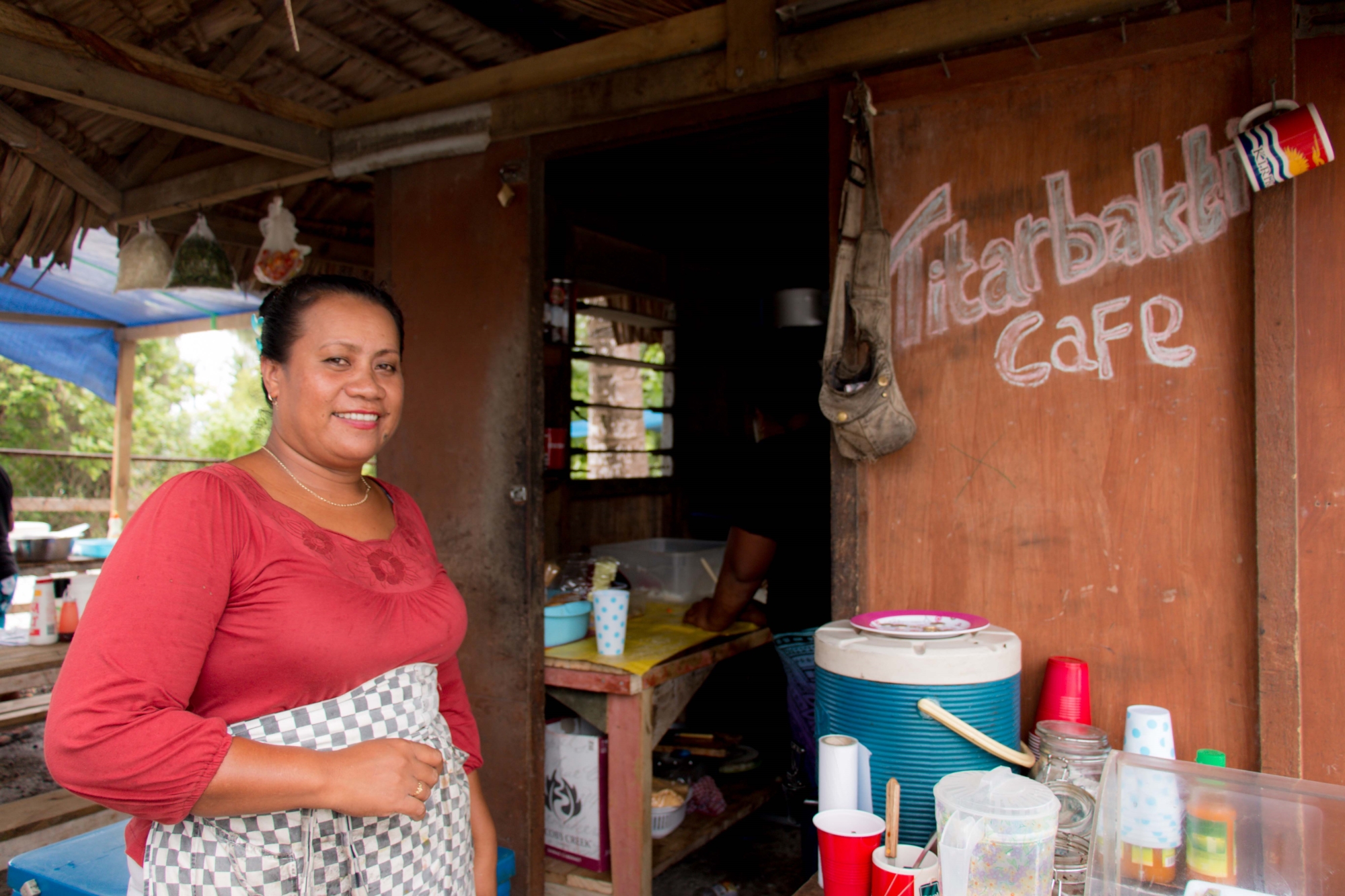 Image of a Kiribati woman wearing a red shirt and a black and white checked apron around her waist. She is standing in front of the cafe she owns, the wall behind her has "Titarbakti cafe" written on it. There is a table in the background with cups, jugs, a water cooler and other containers