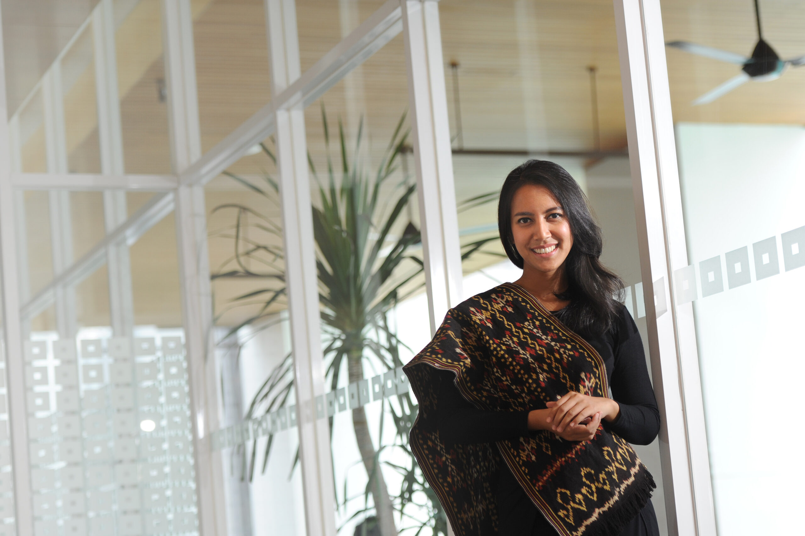 Maryam Rodja, Australia Awards Indonesia awardee is standing in front of large glass windows. She wears a sash with a traditional Indonesian Batik pattern with a black long sleeve top.