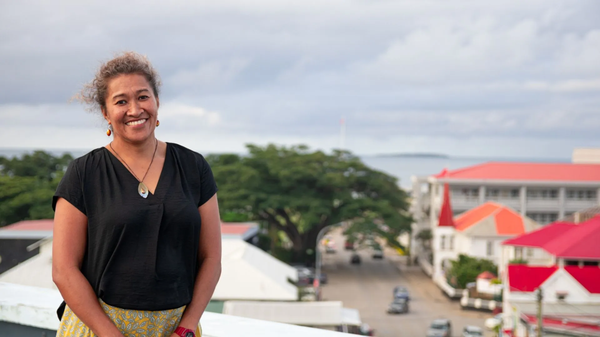 A woman stands smiling against a backdrop of a grey sky, trees and buildings with red and orange roofing