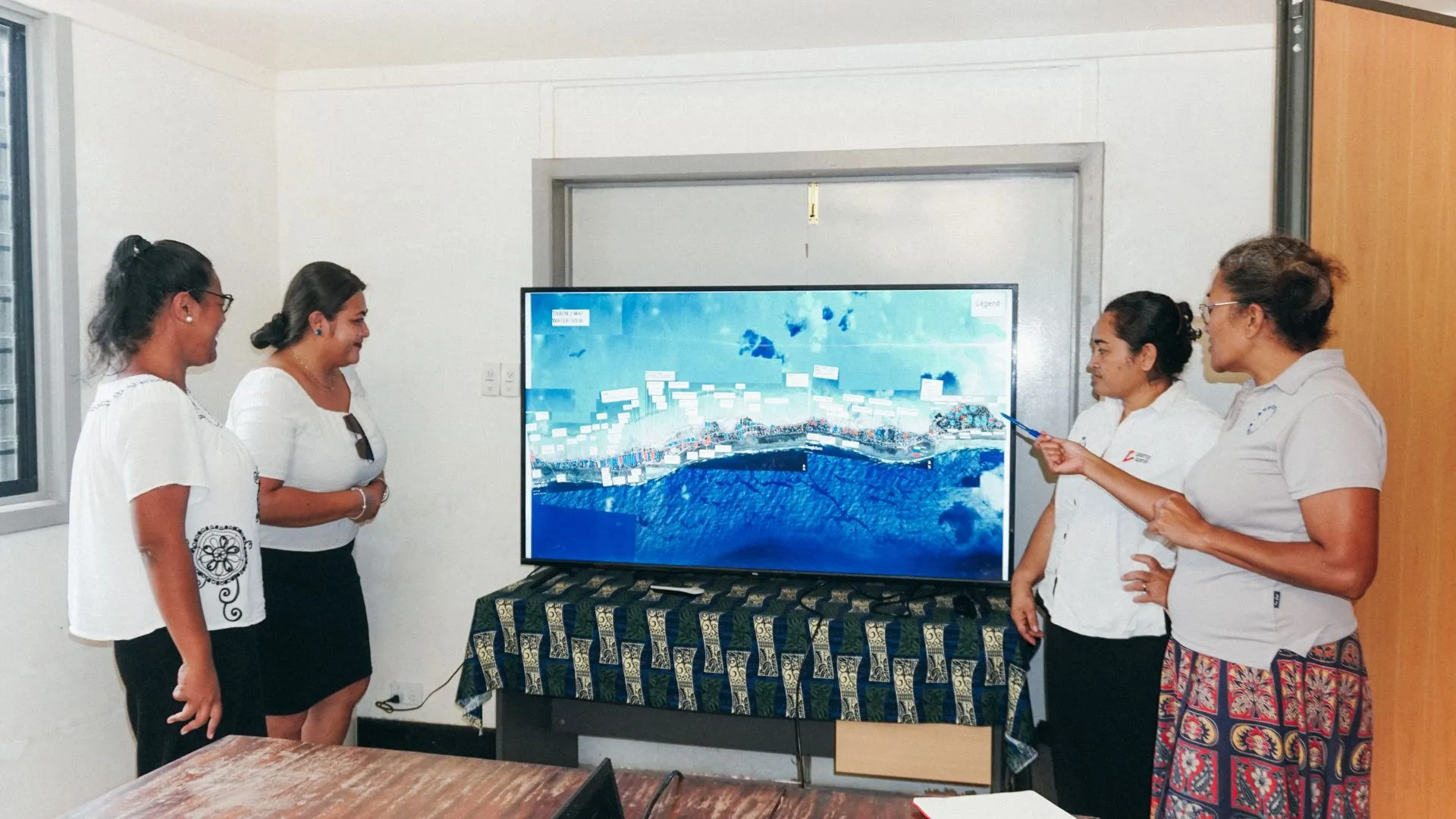 Four women in white t-shirts stand around a large tv screen displaying abstracted blue imagery. 