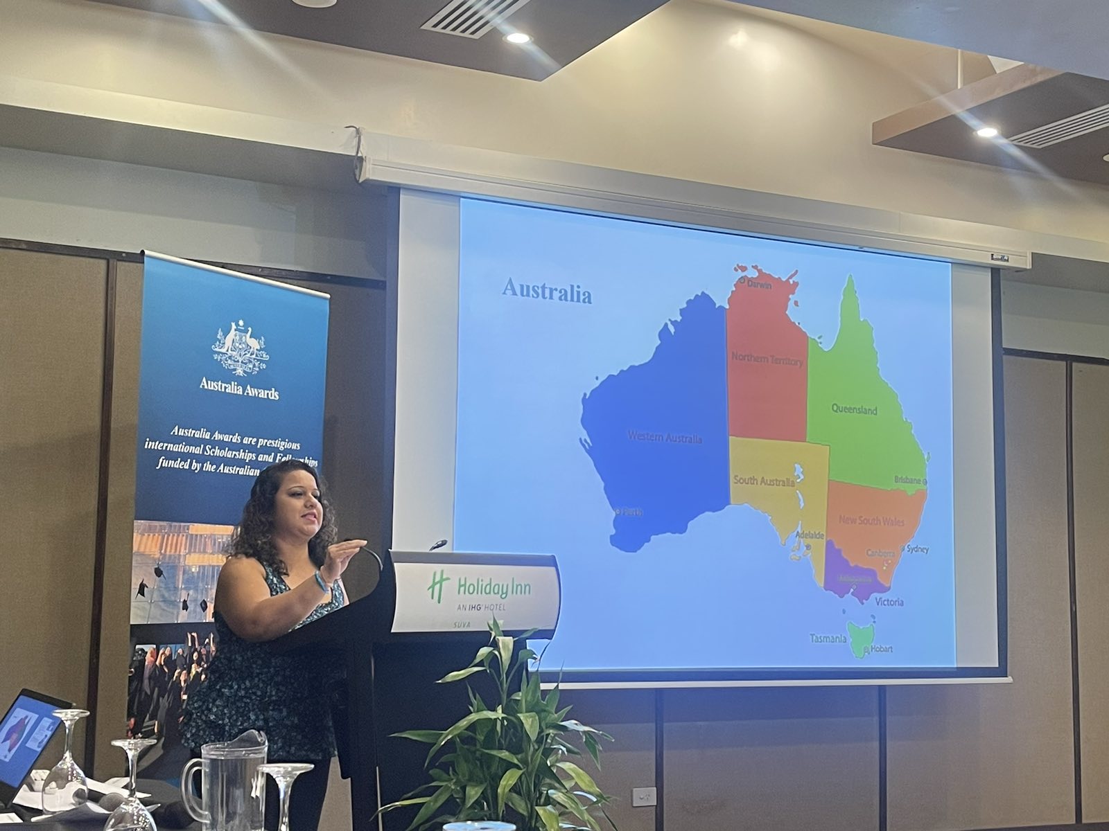 A woman stands in front of a large PowerPoint screen displaying a map of Australia, with states highlighted in different colours