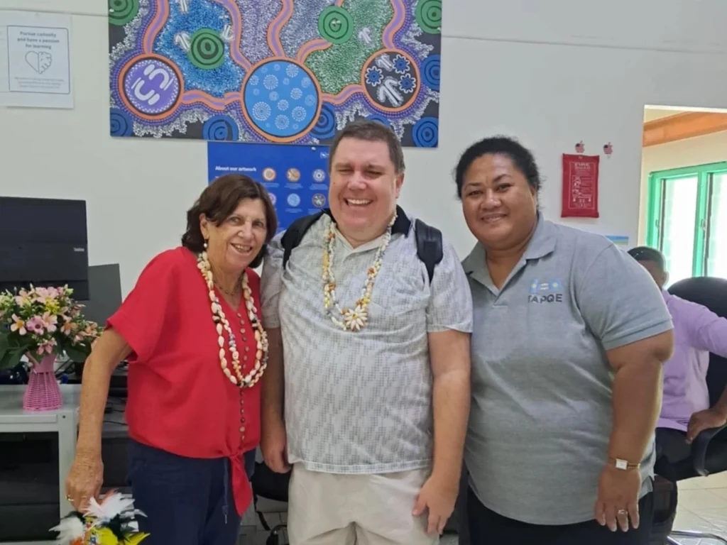 Three people standing in a school room in front of an artwork. On the left is Annie Dares, on the right is Rosie Paueli, and in the middle is Ben Clare. The three are positioned together, with the artwork visible behind them. They appear smiling for the photo. 