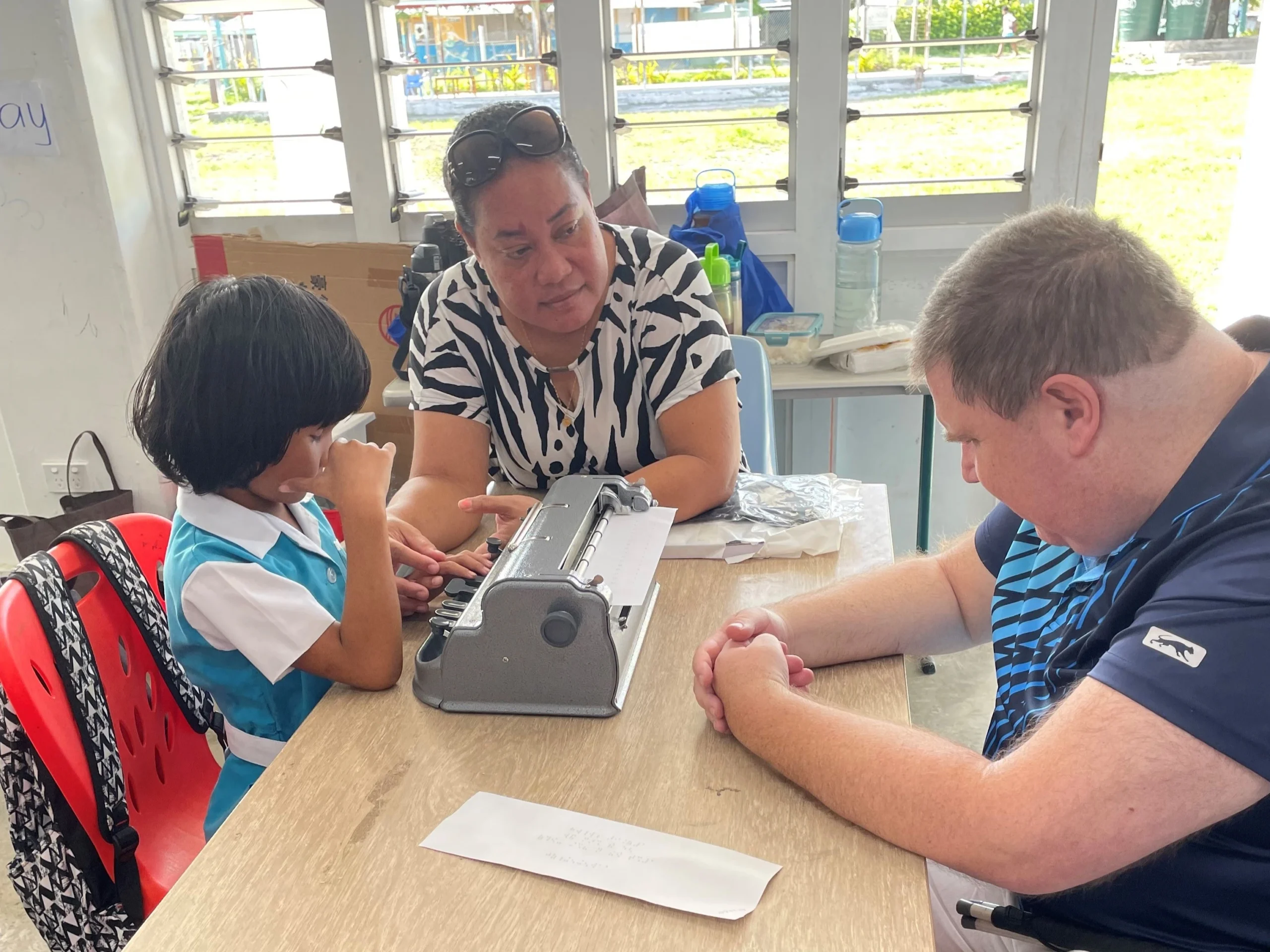 Ben Clare sits at the right side of a small table, opposite seven-year-old Toko, who is using a Braille machine. Toko's teacher, Louise Leitouya, Head Teacher of Aofiaga Centre, sits beside them, at the end of the table. The setting is an educational environment.