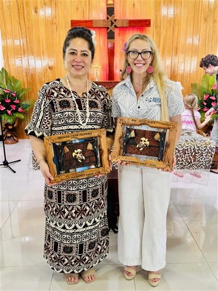 Two women stand side by side smiling for the camera, holding wooden ornamental framed objects. 