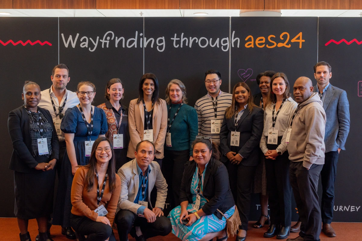 A group of 15 stand in front of a large sign reading "Wayfinding through aes24"